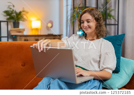 Happy young woman lady sitting on sofa opens laptop and working on project in living room at home 125372185