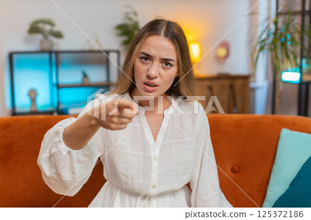 Furious young Caucasian woman sitting on sofa at home, angrily pointing at you through the camera 125372186