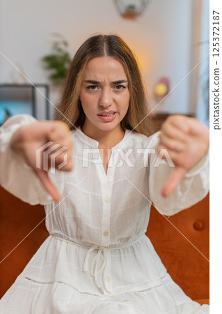 Upset young woman showing thumbs down sign gesture, expressing discontent sitting on sofa at home 125372187