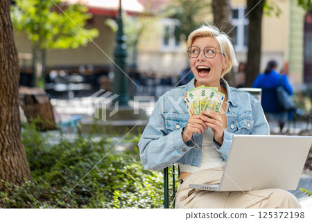 Happy rich young woman using laptop winner victory holding cash money in euro banknotes in city Happy rich young woman using laptop winner victory holding cash money in euro banknotes in city 125372198