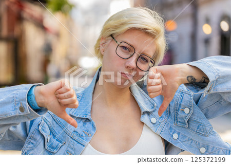 Upset young Caucasian blonde woman showing double thumbs down expressing discontent on city street Upset young Caucasian blonde woman showing double thumbs down expressing discontent on city street 125372199