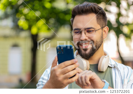 Smiling young man using smartphone typing texting scrolling social media app on city town street Smiling young man using smartphone typing texting scrolling social media app on city town street 125372201