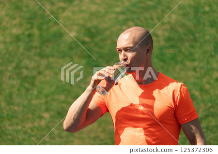Sporty casual man drinking water wearing orange t-shirt on outdoor sports ground. Water balance for heat season for active workout. Close up. 125372306