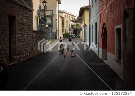 rear view of two small girls riding bicycle and scooter in the street of a town 125372884