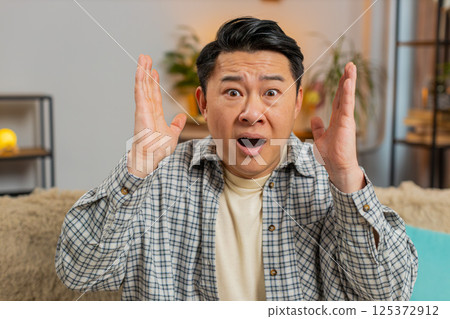 Upset Chinese man making face disappointed in result, bad news watching TV on sofa couch at home Upset Chinese man making face disappointed in result, bad news watching TV on sofa couch at home 125372912