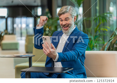 Amazed businessman using smartphone shocked by sudden victory lottery win in modern office lobby 125372914