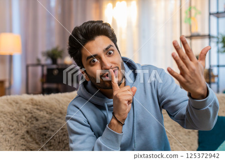 Young Indian man pressing index finger to lips making silence gesture sitting on sofa couch at home Young Indian man pressing index finger to lips making silence gesture sitting on sofa couch at home 125372942