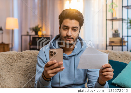 Young Indian man paying utility bills on smartphone with receipts sitting on sofa in living room Young Indian man paying utility bills on smartphone with receipts sitting on sofa in living room 125372943