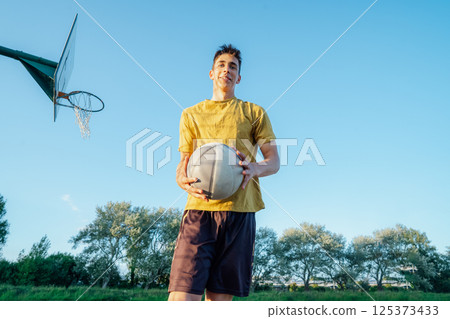 Young caucasian male teenager holding basketball on an outdoor court. Playing Basketball at Park outdoor playground in sunset light. Basketball match. Teenagers' hobby. Sports and healthy lifestyle 125373433