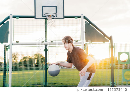 Young male player focuses on dribbling basketball on an outdoor court. Playing Basketball at Park outdoor playground in sunset light. Basketball match. Teenagers' hobby. Sports and healthy lifestyle 125373440