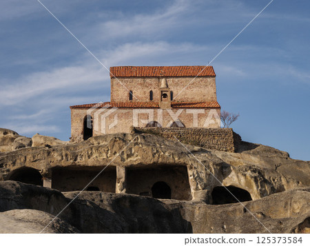 Towering church crowning historic cave complex, revealing ancient Georgian architectural heritage against vibrant blue backdrop Towering church crowning historic cave complex, revealing ancient Georgian architectural heritage against vibrant blue backdrop 125373584