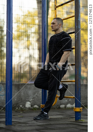 man stands relaxed leaning against fitness bar in outdoor gym as golden autumn sunlight casts warm glow. His casual attire and serene backdrop create calming atmosphere. 125374613