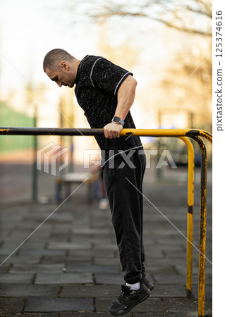 man is engaged in strength training routine outdoor gym demonstrating endurance and fitness he performs dip exercise on bar surrounded lush trees and clear sky. 125374616