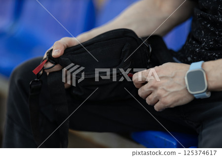 person sits comfortably on blue bleachers in indoor area focused on their small black bag and adjusting zipper while wearing casual wristwatch and relaxed clothing. 125374617