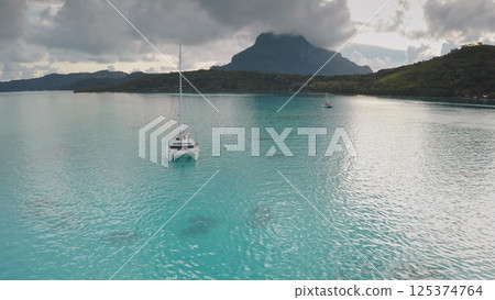 Bora Bora discovery and travel: white catamaran sailing in tranquil turquoise water with mount Otemanu in background under cloudy sky, creating serene tropical paradise view. Remote wild nature scene Bora Bora discovery and travel: white catamaran sailing in tranquil turquoise water with mount Otemanu in background under cloudy sky, creating serene tropical paradise view. Remote wild nature scene 125374764