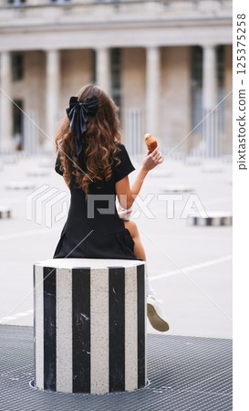 Relaxed girl with curly hair enjoys croissant and coffee in French setting 125375258