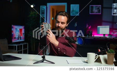 Tech guru presenting V mount battery, filming technology review using cellphone on tripod and professional microphone. Caucasian vlogger showcases charging device specs, recording with phone, camera B 125375598