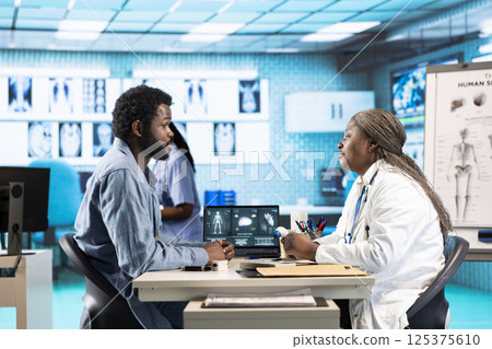 Medical professional explaining the x ray imaging for a diagnosis, doing scan interpretation during check up visit. African american female medic gives feedback to male client. 125375610