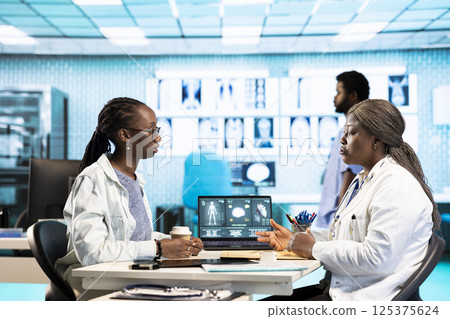 African american medic practitioner discuss with a girl about health care concerns, utilizing medical data and tech equipment to enhance treatment and prevention. Clinic examination. 125375624