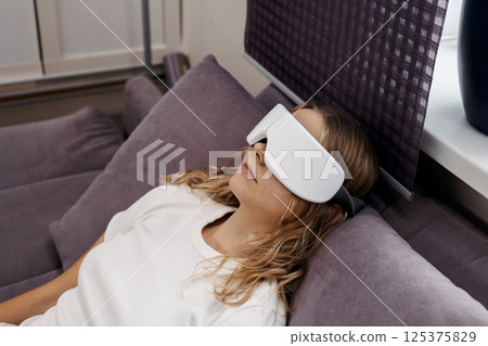Eye massage device. Young girl wearing glasses for massage relaxing on sofa at home. Woman takes care of her health and skin care 125375829
