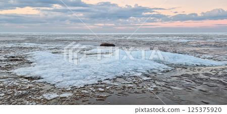 Sunrise over a tranquil coastline with icy patches and smooth stones along the sandy shore. The sky reflects in the water, creating a serene and peaceful atmosphere. 125375920