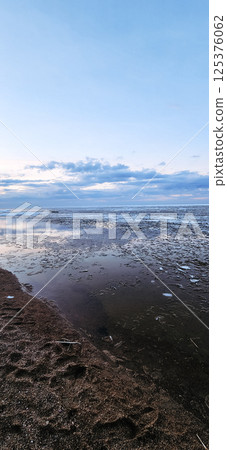 Spring landscape. Panoramic view of beautiful dawn on bay. Cumulus clouds over water in bright light. Ice, snow and rocks on coastline. Rising rays of sun are reflected in sea. Vertical photo. 125376062