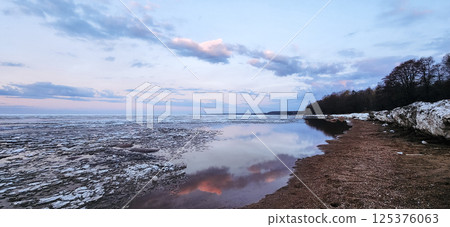 Spring landscape. Panoramic view of beautiful dawn on bay. Cumulus clouds over water in bright light. Ice, snow and rocks on coastline. Rising rays of sun are reflected in sea. High quality photo 125376063