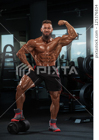 muscular individual poses proudly in fitness center emphasizing his sculpted physique. He stands on one leg showcasing his strength while surrounded gym equipment. muscular individual poses proudly in fitness center emphasizing his sculpted physique. He stands on one leg showcasing his strength while surrounded gym equipment. 125376834