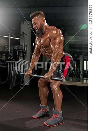 well built man in fitness center performs intense workout cable machine designed build upper body strength and enhance muscle definition. Bright lighting emphasizes his hard work. 125376835
