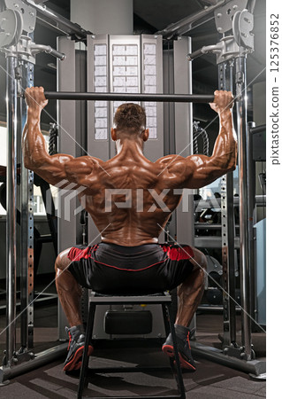 well built man showcases strength as he performs seated overhead press using weight machine in modern fitness studio during daylight. His muscles are prominently defined. well built man showcases strength as he performs seated overhead press using weight machine in modern fitness studio during daylight. His muscles are prominently defined. 125376852