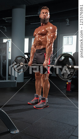 muscular man performs deadlift barbell in well equipped gym. He focuses on strength training showcasing dedication fitness. Natural light filters through large windows. 125376861