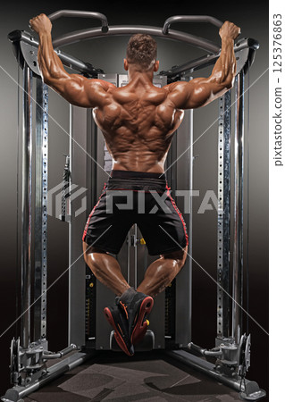 well built man performs pull ups on gym equipment strengthen his back and arms. environment is focused on fitness showcasing dedication health and strength training. well built man performs pull ups on gym equipment strengthen his back and arms. environment is focused on fitness showcasing dedication health and strength training. 125376863