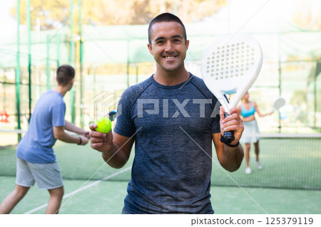 Portrait of a satisfied adult man with racket and ball in his hands on padel court Portrait of a satisfied adult man with racket and ball in his hands on padel court 125379119