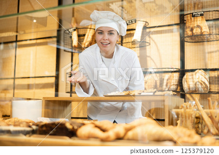 Smiling young saleswoman in white chef jacket and cap standing behind bakery counter during working day in bakehouse. Cooking, culinary and bakery concept Smiling young saleswoman in white chef jacket and cap standing behind bakery counter during working day in bakehouse. Cooking, culinary and bakery concept 125379120