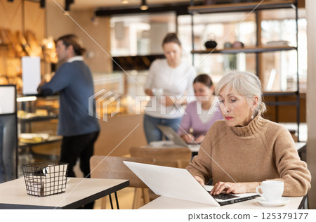 Positive European old woman drinking and enjoying coffee while working on laptop in cafe 125379177