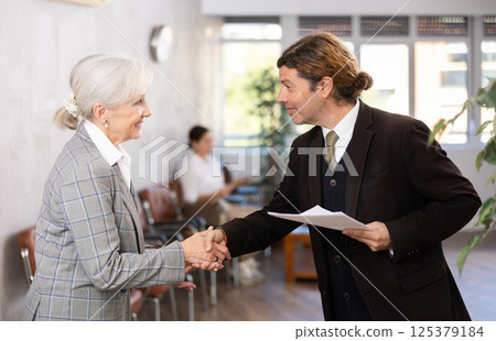 Smiling businessman with papers shaking hands with elderly woman Smiling businessman with papers shaking hands with elderly woman 125379184