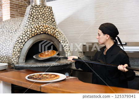 Female chef in black uniform prepares pizza and pulls it out of oven in kitchen of pizzeria Female chef in black uniform prepares pizza and pulls it out of oven in kitchen of pizzeria 125379341
