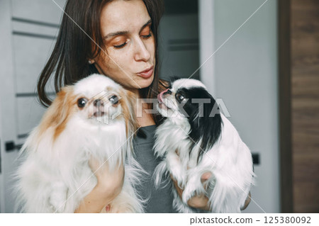 woman tenderly holding two japanese chin dogs, showcasing affection and pet care at home 125380092