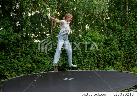 woman jumping on trampoline in jeans and white shirt near lush green forest woman jumping on trampoline in jeans and white shirt near lush green forest 125380216