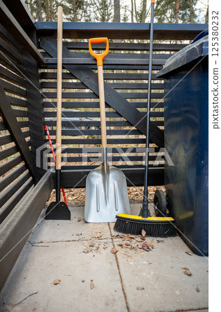 Various garden tools including broom, shovel, and hoe leaning against a wooden fence outdoors Various garden tools including broom, shovel, and hoe leaning against a wooden fence outdoors 125380232