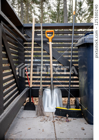 Various garden tools including broom, shovel, and hoe leaning against a wooden fence outdoors 125380240