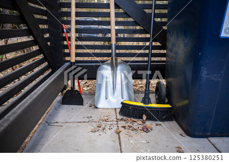 Various garden tools including broom, shovel, and hoe leaning against a wooden fence outdoors Various garden tools including broom, shovel, and hoe leaning against a wooden fence outdoors 125380251