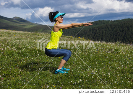 Woman doing squats at flowering grassland mountain top Woman doing squats at flowering grassland mountain top 125381456
