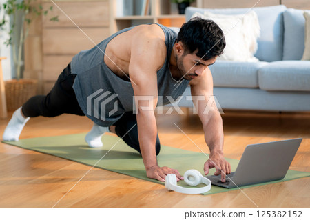 Young asian man doing home workout while watching online tutorial on laptop computer in living room. 125382152