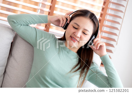 Happy young asian woman wearing headphones listening music while sitting on sofa with fun. 125382201