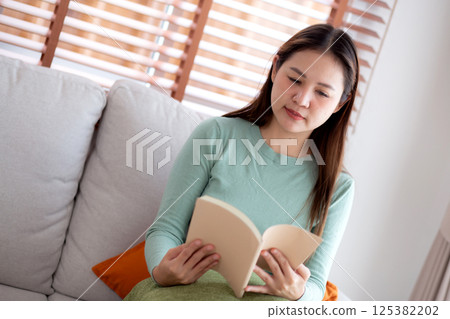 Young asian woman reading a book with expression sitting on sofa in living room at home. 125382202