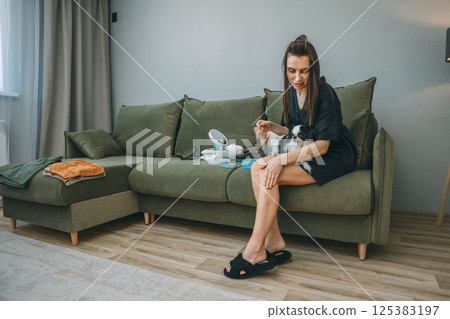woman applying skincare lotion to her leg while sitting on green couch with two japanese chin dogs during relaxed morning beauty routine, pets calm, skincare, dog lovers, self care brands 125383197
