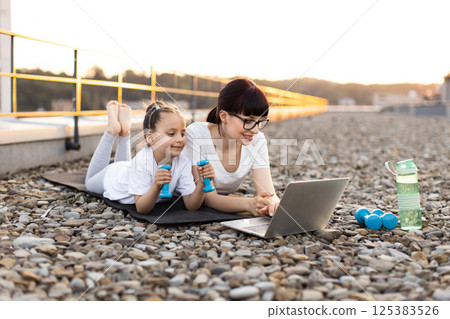 Adult woman and young girl lying outside learning exercise techniques from laptop together. Concept of bond, fitness, outdoor activity. Dumbbells and bottle on mat indicating active session. 125383526