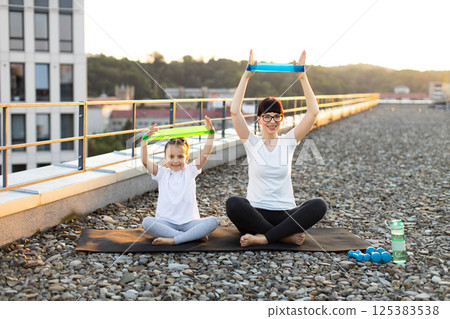 Adult woman and young girl performing fitness exercises with resistance bands on rooftop outdoors. Creative family workout promoting healthy lifestyle and physical fitness Adult woman and young girl performing fitness exercises with resistance bands on rooftop outdoors. Creative family workout promoting healthy lifestyle and physical fitness 125383538