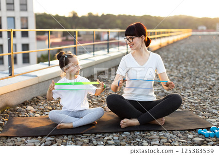 Mother and young daughter sitting and stretching resistance bands outdoors on rooftop during sunset. Emphasizing family bonding, fitness, active lifestyle, and healthy habits. Mother and young daughter sitting and stretching resistance bands outdoors on rooftop during sunset. Emphasizing family bonding, fitness, active lifestyle, and healthy habits. 125383539